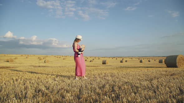 Stylish Mother Lulling Baby Son in Hands in Hay Field During Hot Summer Day alt