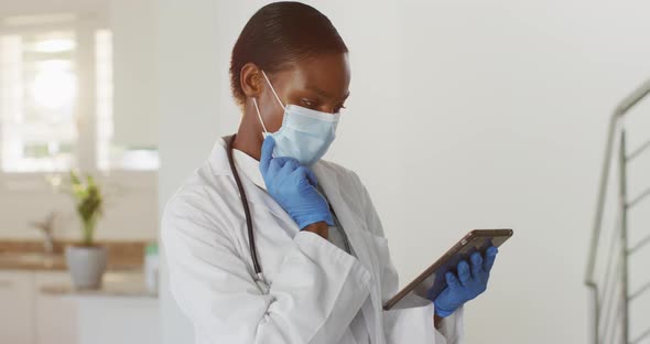 African american female doctor wearing mask having video call consultation using digital tablet alt