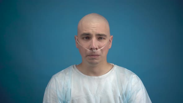 A Bald Young Man with Oncology Looks at the Camera and Cries on a Blue Background. The Patient alt