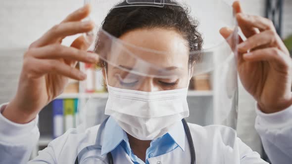 Doctor Woman in Protective Mask is Putting on a Face Shield and Looking at you While Standing in alt