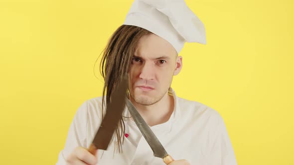 Young Serious Man Dressed As Chef with Knives on Yellow Background alt