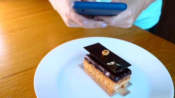 Woman Hand with Mobile Phone Take Picture of Opera Cake Dessert on White Plate alt