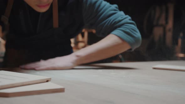 Male Carpenter a Young Man in Work Clothes and an Apron Wipes Dust and Sawdust From the Work Table alt