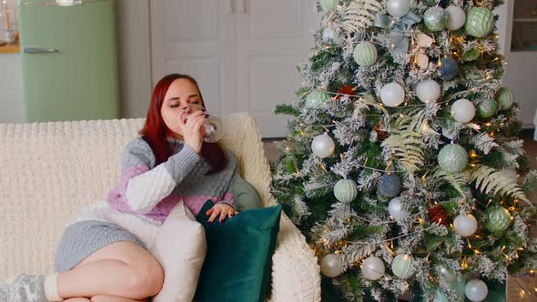 Woman Sitting on Sofa Near Christmas Tree and Drinking Alcoholic Beverage alt
