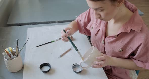 Female Potter Sitting and Stirs Paint with a Brush a Cup on the Table. Woman Making Ceramic Item