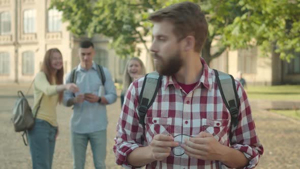 Nervous Bearded University Student Looking Back at Groupmates Laughing and Pointing at Him, and alt