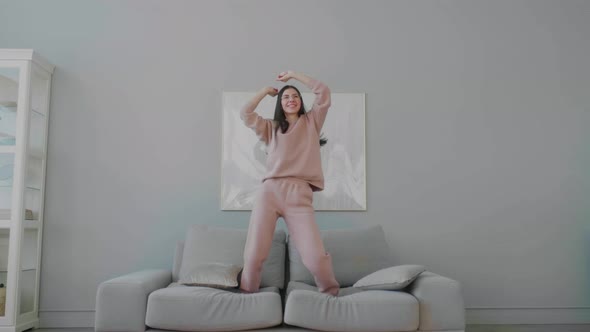 Young caucasian woman wearing glasses having fun, dancing and jumping on the sofa, waving her arms.