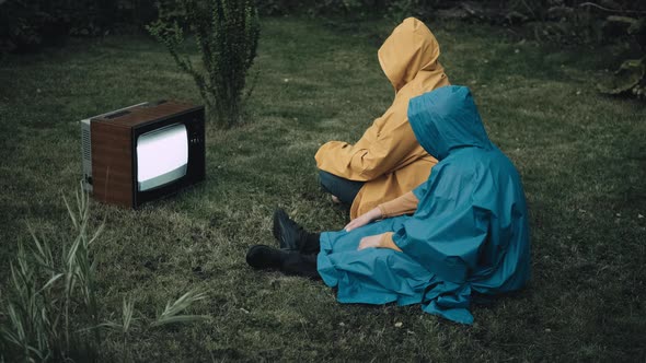 Man and Woman in Raincoats with Hood are Sitting on Grass and Watching an Old TV