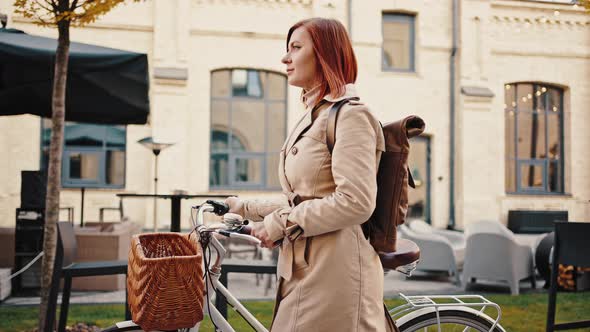 Young Redhead Female is Riding Cruiser Bike By Deserted City Square Decorated with Lights alt