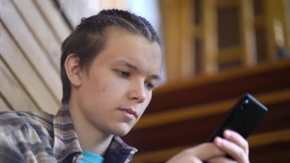 Teen Boy Sitting on Stairs at Home and Using App on His Mobile Phone alt