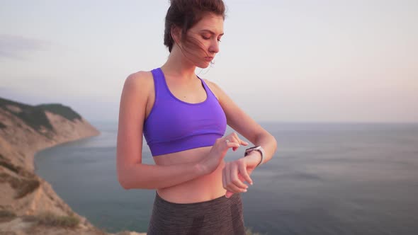 Young Beautiful Girl in Sportswear Is Touching a Smart Watch at Sunset By the Ocean