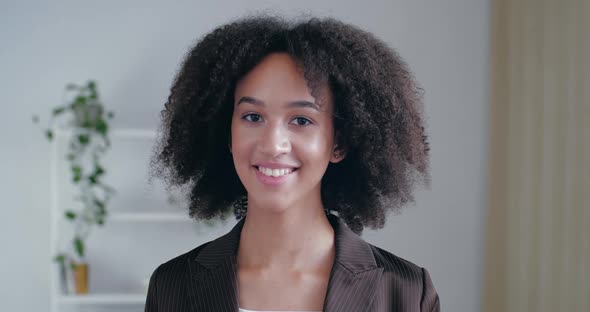 Сurly-haired Young American Woman Stands in Office at Home in Room in Front of Camera, Smiles alt