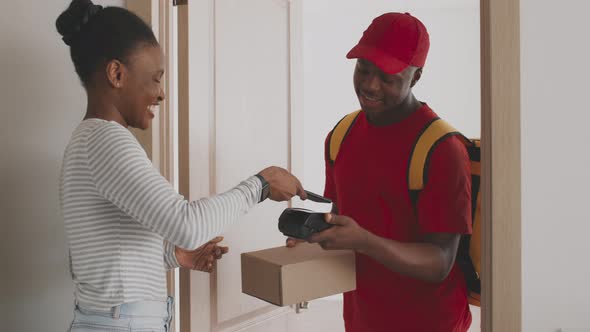 Young African American Woman Paying for Order with Smartphone and Getting Package Box From Postal alt