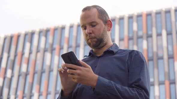 A Caucasian Man Works on a Smartphone  Closeup From Below  an Office Building in the Background