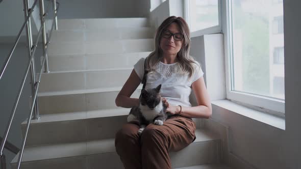 Pretty Woman Strokes Colorful Stray Cat While Sitting on White Steps of Staircase of Apartment alt