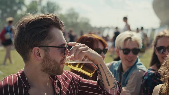 Group of friends sitting on grass together at music festival and drinking beer while chatting. Shot alt