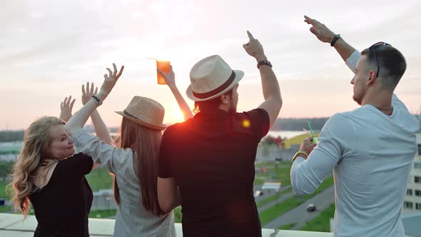 Rear View Group of People Enjoying Beautiful City Landscape Raising Arms on Rooftop Party at Sunset alt