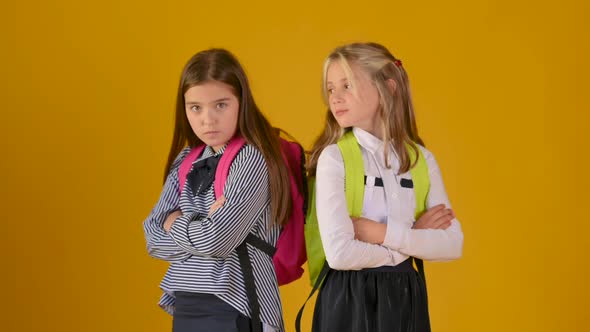 Two schoolgirls in school uniforms with backpacks resentfully look at each other alt