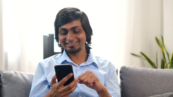 Serene Smiling Young Indian Man Sitting Down on the Sofa at Home with a Smartphone alt