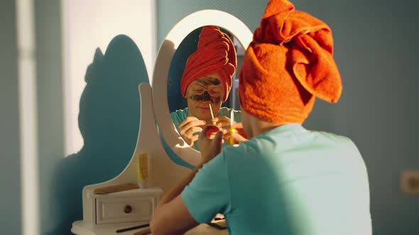 Young man in a cosmetic mask with a towel on his head looks at himself in the mirror alt