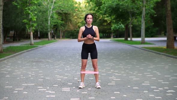 Young Woman Athlete Exersices with Resistance Band Doing Squats Outdoors at Local Green Public Park alt