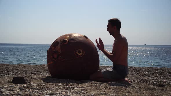 Man Doing Yoga Exercise Outdoors Near to an Old Rusty Floating Marine Mine on the Beach with Rocky alt