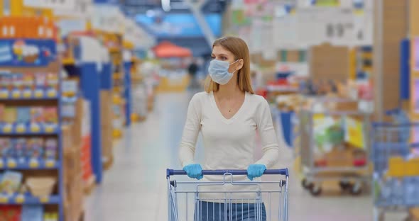 Portrait Beautiful Girl in a Medical Mask Goes to the Supermarket with a Trolley and Looks Around alt