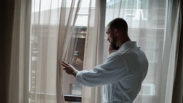 a Man in a White Coat Stands at the Window Holds a Phone to Ear Peeks Through the Curtain and Lets alt