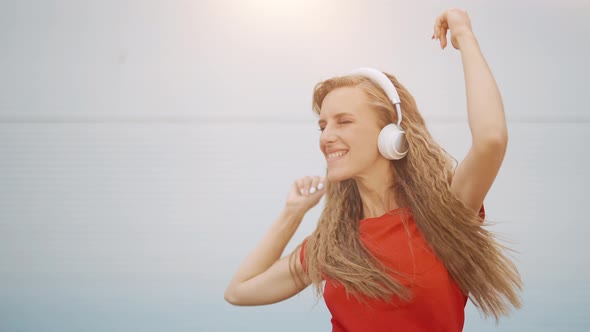A Young Girl in Headphones is Listening to Music and Amusingly Dancing on a Light Background alt