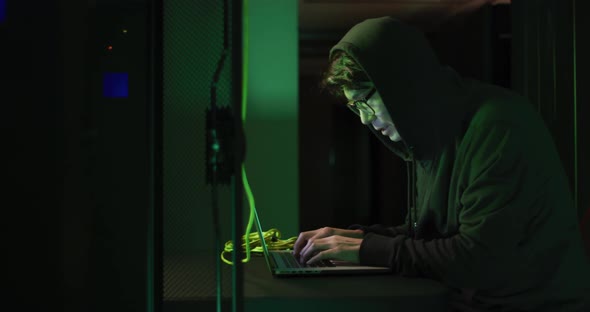 Asian male hacker in hoodie using laptop by computer servers alt