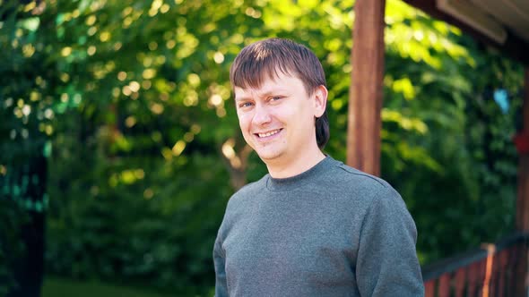 Close-up, Portrait: Young Man Stands on the Background of a Summer House and Greenery and Smiles alt