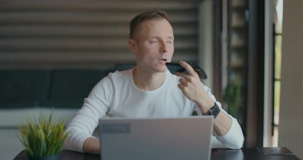 Young Man Sits in Room at Laptop and Records Voice Message on His Smartphone alt