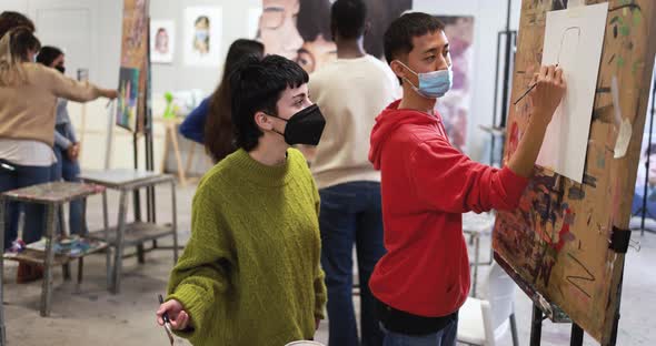Multiracial students painting inside art room class wearing safety masks alt