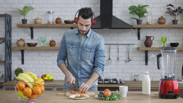 Handsome Joyful Middle Eastern Man in Headphone Cutting Tomato for Healthful Vegan Salad and Dancing alt