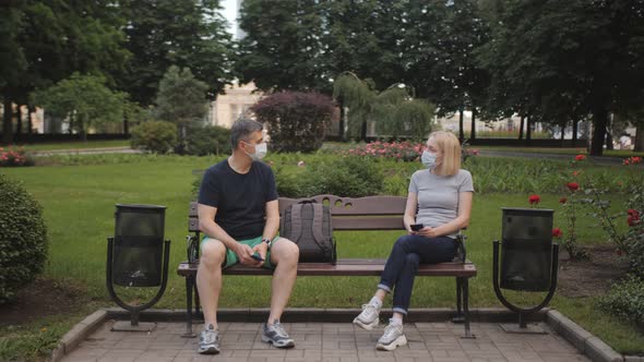 A Man and a Woman in Protective Masks Sit at Opposite End of a Bench and Use Phones in the Park