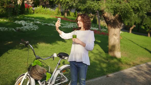 Portrait of a Young Woman Taking Selfie Photo with Cellphone Standing in the City Park Near Her City alt