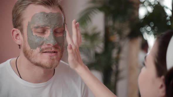 Woman Applying Beauty Mask on Husband Boyfriend Face. Girlfriend Taking Care Cleansing Skin