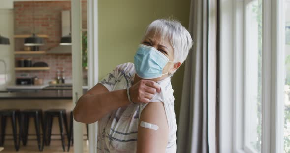 Happy mixed race senior woman in face mask showing plaster on arm after covid vaccination alt