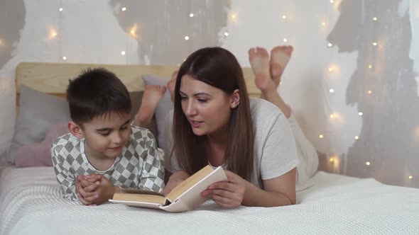 Mum Reads a Book to Her Son Lying on a Bed in Pajamas and Kisses Him