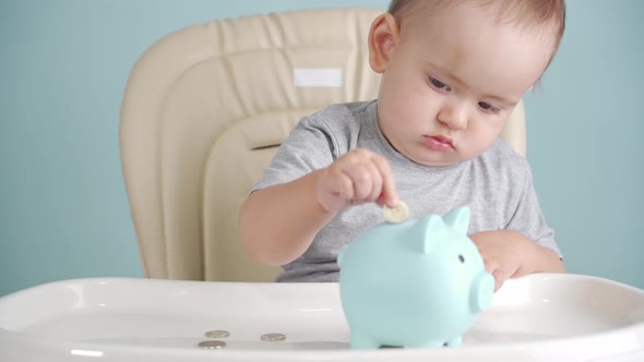 Toddler 12-17 months old throws a coin into a blue piggy bank sitting on a chair, selective focus alt