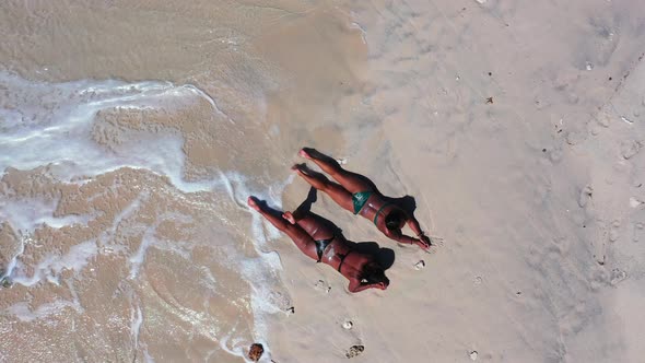 Two girls in bikini with tanned skin lying on white sand of exotic beach washed by white waves foami alt