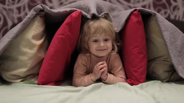 Little Girl Hides Under Blanket and Pillows on the Bed. Concept of Hiding Place alt