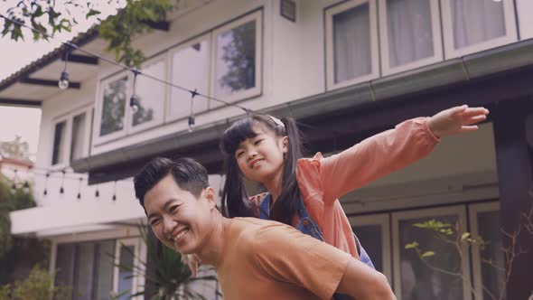 Young Asian Father Giving His Daughter Piggyback and Smiling in Front of House alt