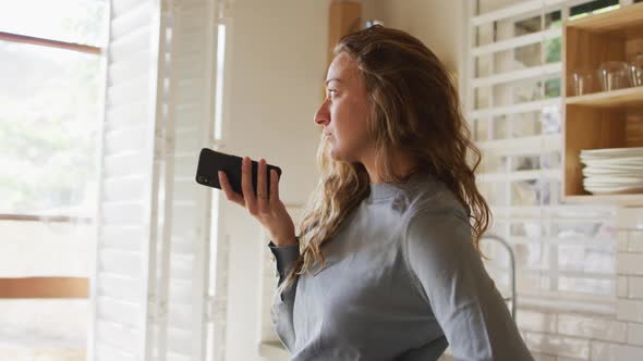 Thoughtful caucasian woman in sunny cottage kitchen holding smartphone looking out of window alt