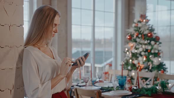 Blonde Woman is Texting on Phone in Front of Dining Table and Christmas Tree alt