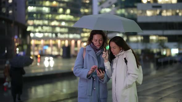 Two Women are Standing Under an Umbrella Looking at the Phone and Laughing Against the Background of alt