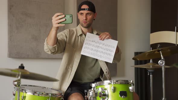 Young Man Records Drum Lesson on Phone Showing Music Sheet alt