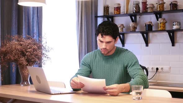 Man Working on Laptop and Reading Documents in Kitchen Paperwork alt