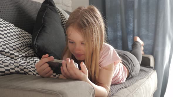 Little Girl Is Lying on the Bed and Looking Into Her Smartphone alt