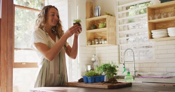 Smiling caucasian woman tending to potted plants standing in sunny cottage kitchen alt
